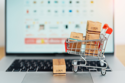 cart The image shows a miniature shopping cart filled with small cardboard boxes, placed on a laptop keyboard. In the background, the laptop screen displays a blurred online shopping site.