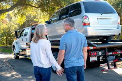street scene The image depicts a couple holding hands and standing in front of a tow truck that has a silver SUV loaded onto it. They are outdoors, with trees and sunlight in the background.